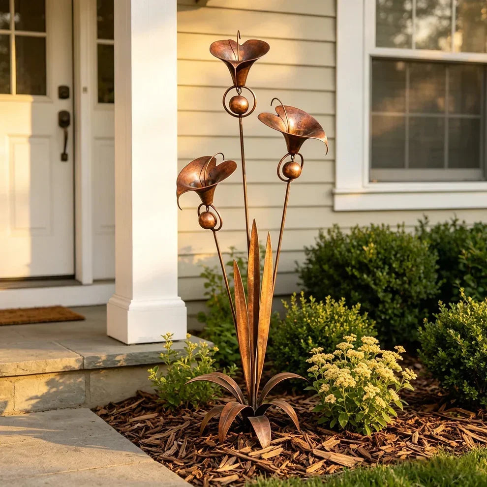 Copper trumpet-flower garden stake standing in wood-chip mulch beside a white porch column, with small yellow flowers nearby. 2