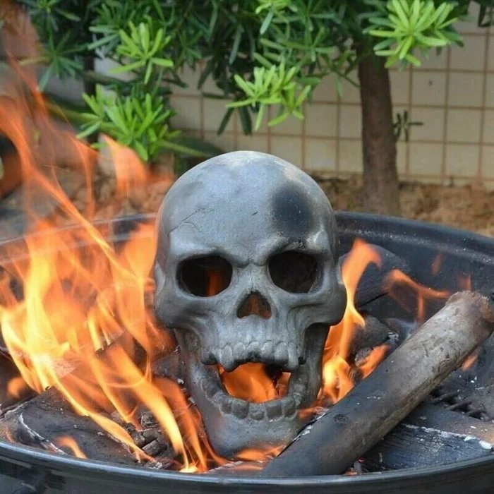 Another view of a single gray skull in a round backyard fire bowl with flames beside a charred log.