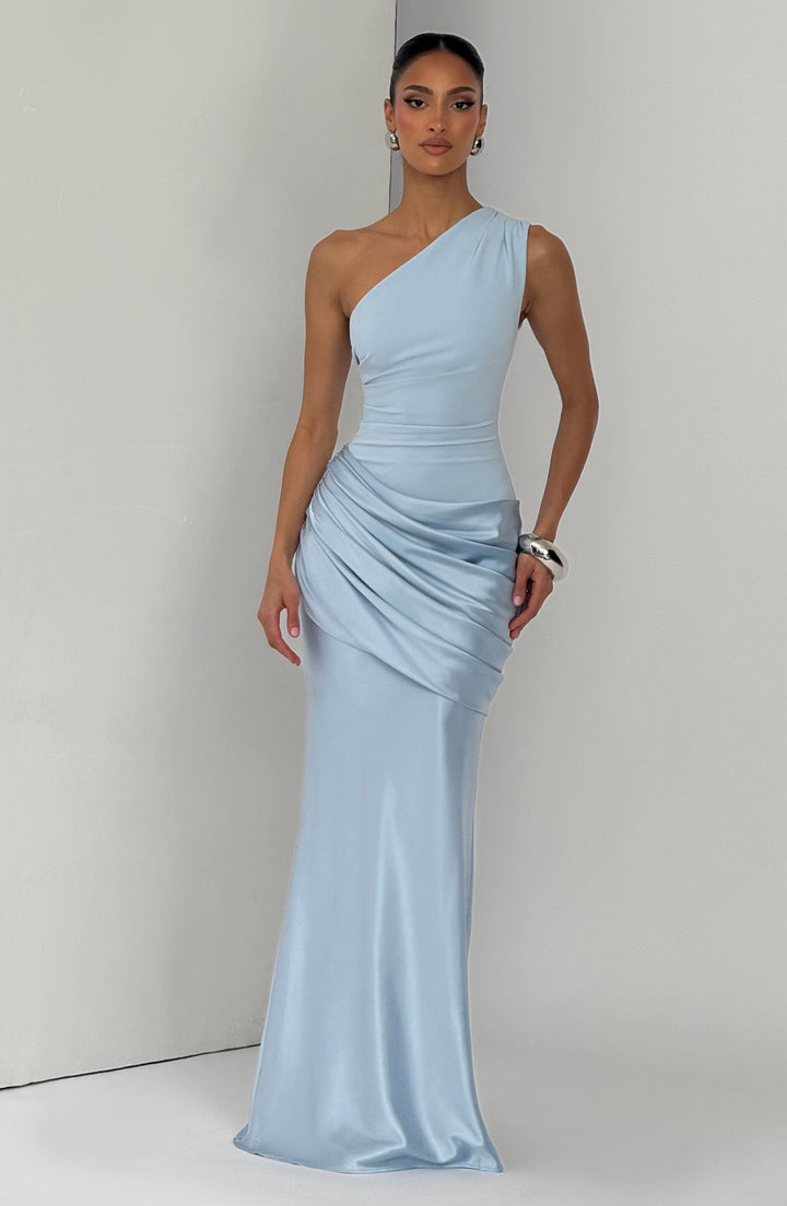 Light blue one-shoulder gown photographed against a pale studio corner, with the model's hand resting on the draped hip. 6
