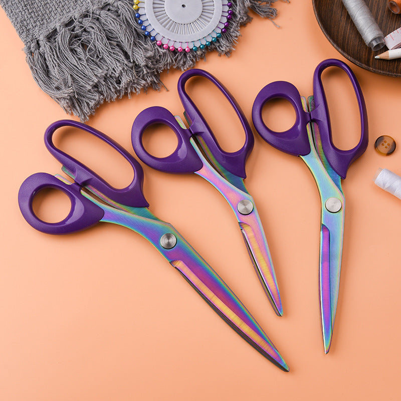Three sewing scissors with purple handles and rainbow-sheen blades laid on an orange surface among pins, thread and fabric.