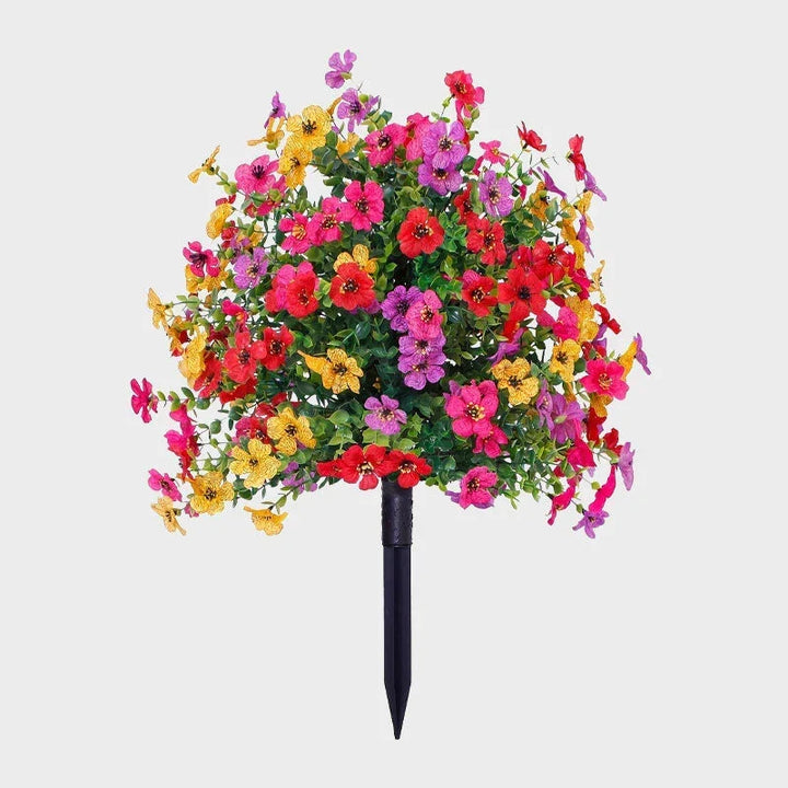 Multicolor artificial flower bush with green foliage on a black ground stake against a plain background.