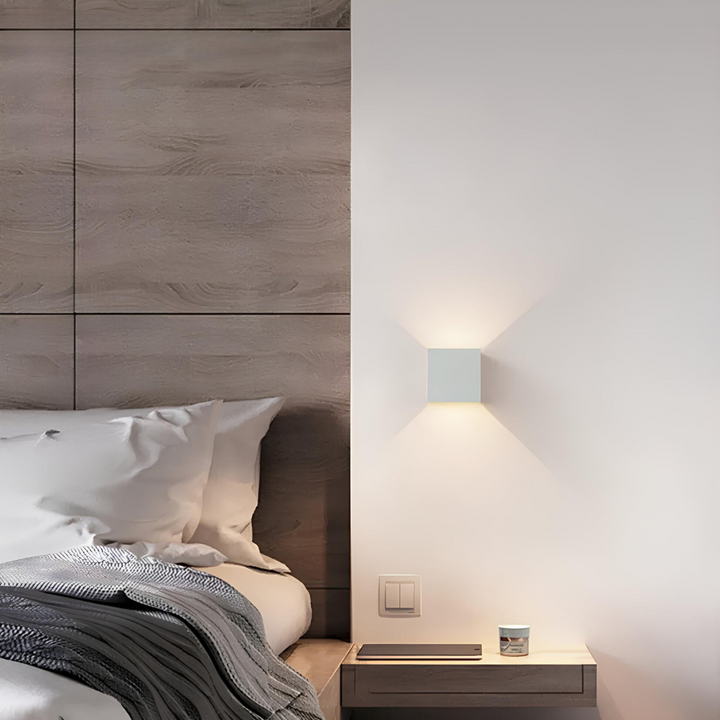 White cube wall lamp mounted beside a bed with a wood-paneled headboard, shown in a bedroom with a floating nightstand. 6
