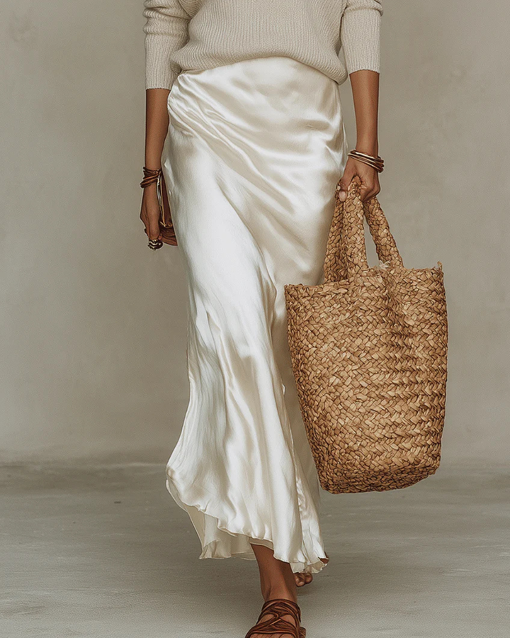 Lower-body view of the flowing ivory satin midi dress with a draped hem, worn with strappy sandals and a woven straw tote. 10