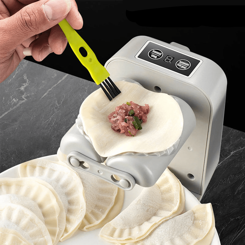 A hand uses a small green-handled brush to wet the edge of a dough round holding minced filling on the machine, sealed dumplings on a plate below. 1