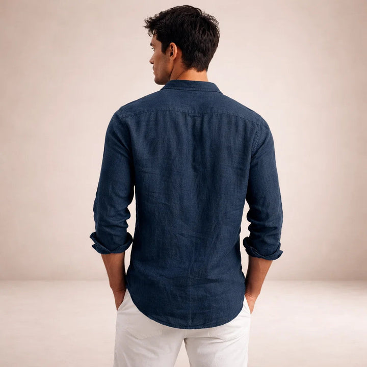 Back view of the navy linen shirt on a model with hands in white pants pockets, showing the yoke seam and rolled sleeves.
