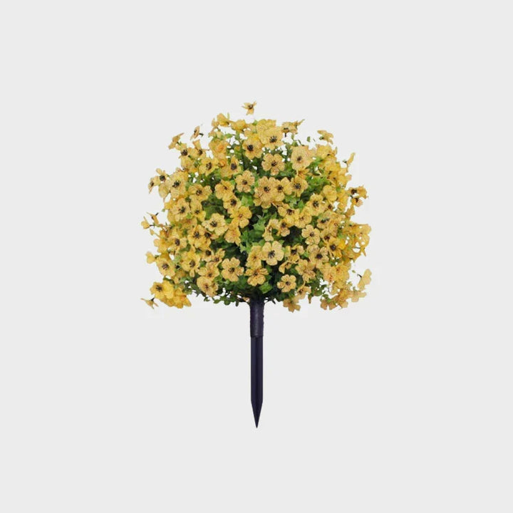 Yellow artificial flower bush with green leaves on a black ground stake against a plain background.