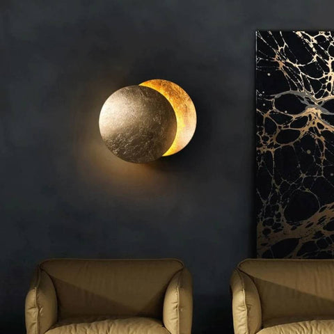 Gold disc wall lamp on a dark wall beside marbled art, above two olive armchairs 3