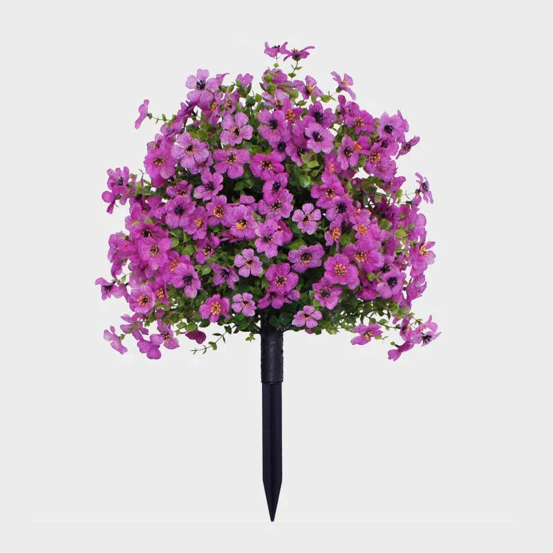 Purple artificial flower bush with green leaves on a black ground stake against a plain background.