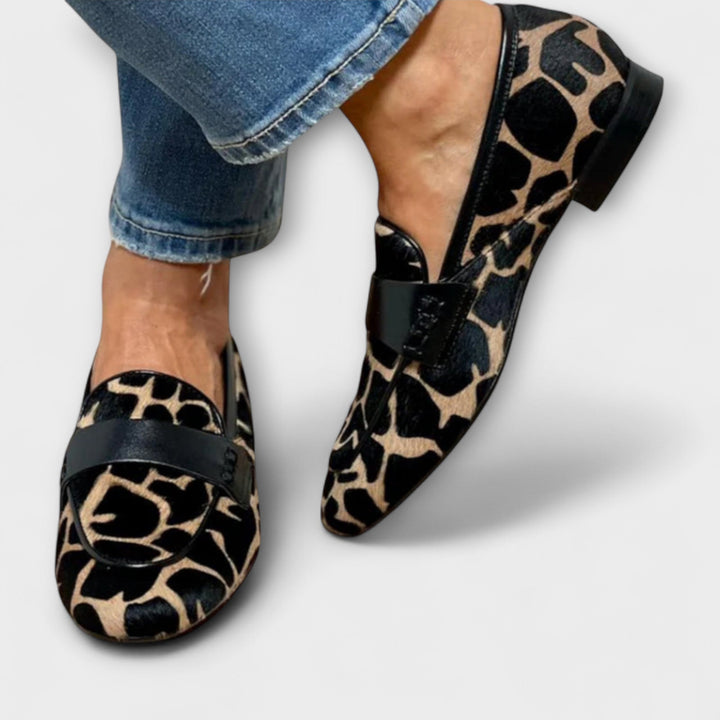Both loafers worn from the front, showing the rounded toe, black strap across the vamp, black piping and low black heel. 3