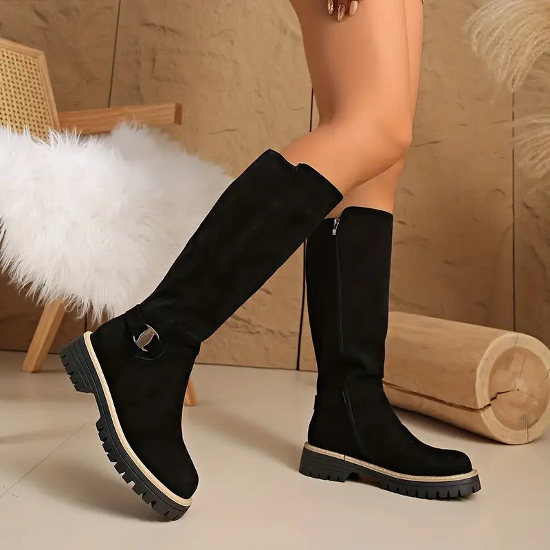 Black knee-high boots with a metal buckle strap and chunky lug sole, standing beside a wooden log.