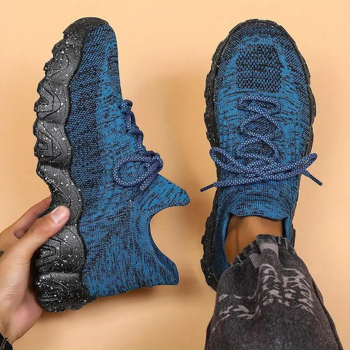 Top-down view of blue marled knit lace-up sneakers with black speckled soles, one held in hand and one worn with gray pants. 28
