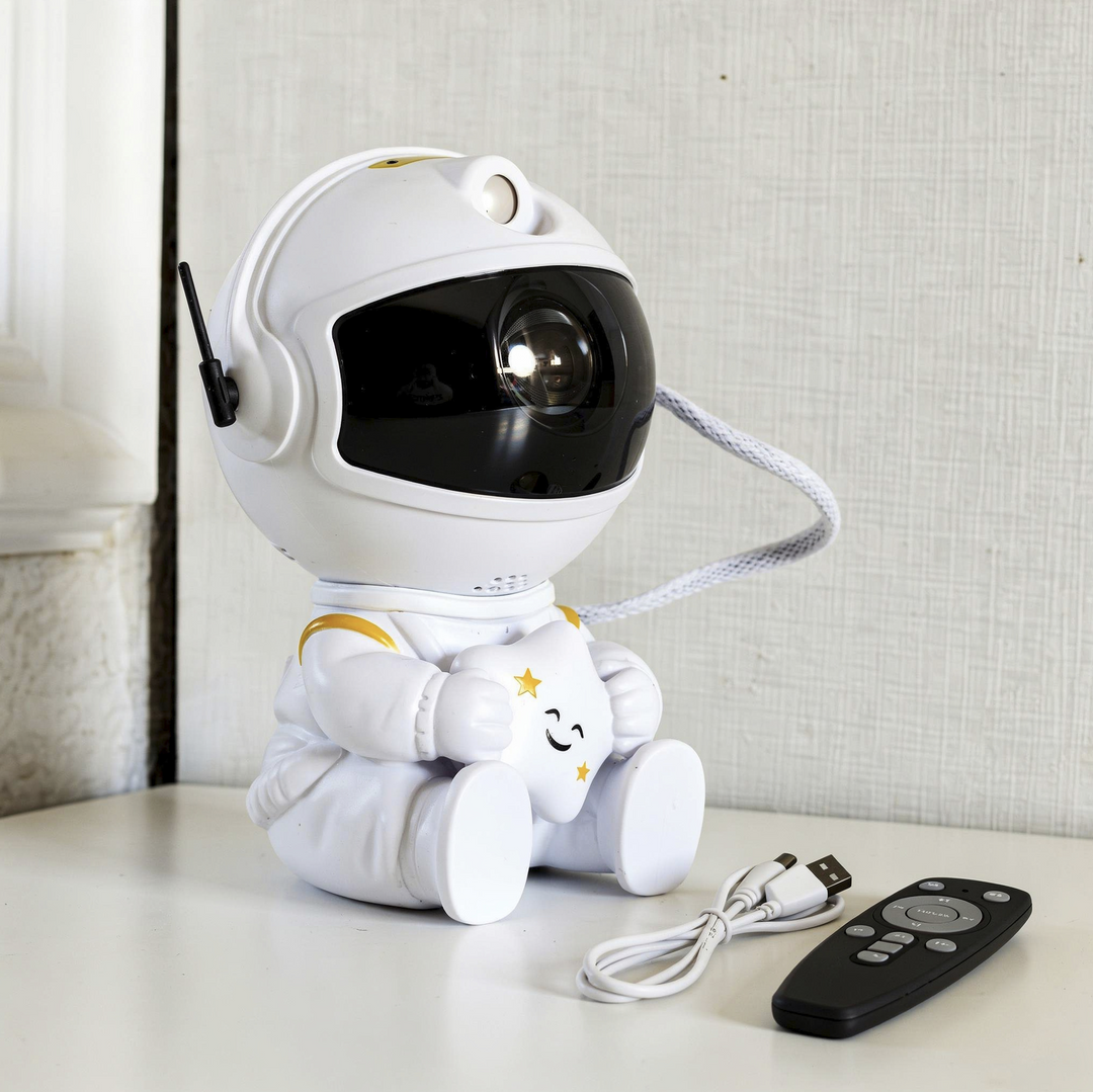 White astronaut projector holding a smiling star, shown from an angle with a USB charging cable and a black remote beside it.