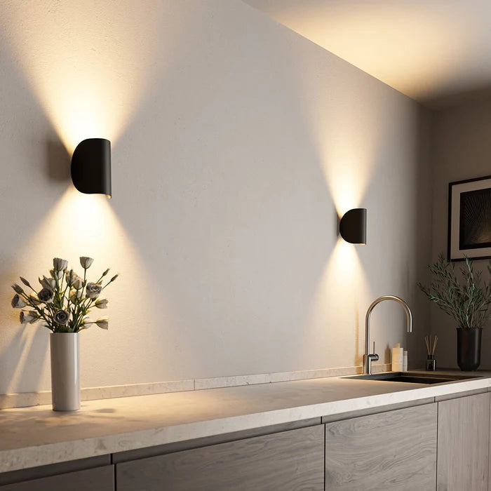 Two black metal LED wall lights on a kitchen wall casting warm light up and down above a counter.