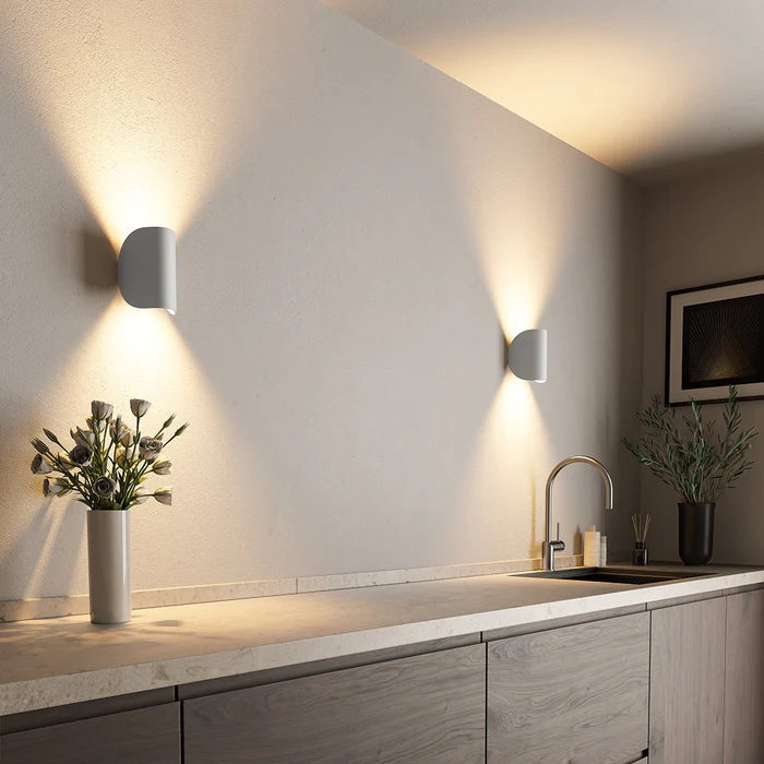 Two white wall lights on a kitchen wall casting warm up and down light over a counter with flowers.