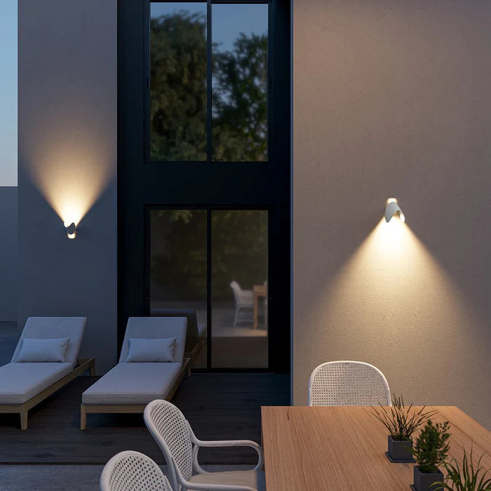 Two white wall lights tilted on exterior walls of a patio at dusk, lighting loungers and a wooden dining table.