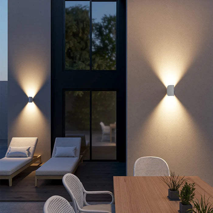 Two white wall lights mounted upright on patio walls at dusk, casting light up and down beside lounge chairs and dining table.