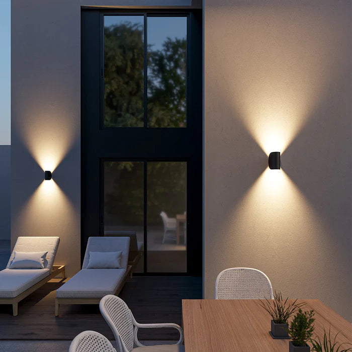 Black wall lights mounted on patio walls at dusk, casting warm light up and down near lounge chairs and a dining table.