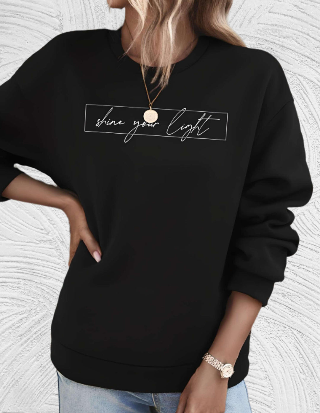 Frau in schwarzem Pullover mit "shine your light" Aufdruck, lässiger Stil, goldene Uhr.