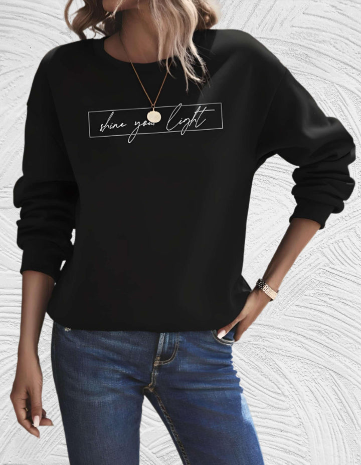 Frau in schwarzem Sweatshirt mit "shine your light" Aufdruck, blaue Jeans, lässiger Stil.