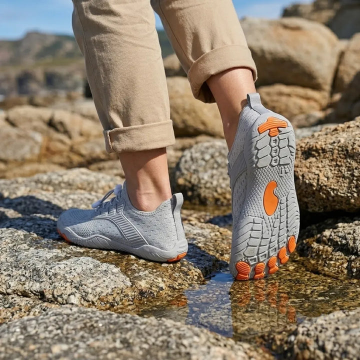 Model in rolled khaki pants wearing gray barefoot sneakers on wet rocks, one sole lifted to show orange tread accents.