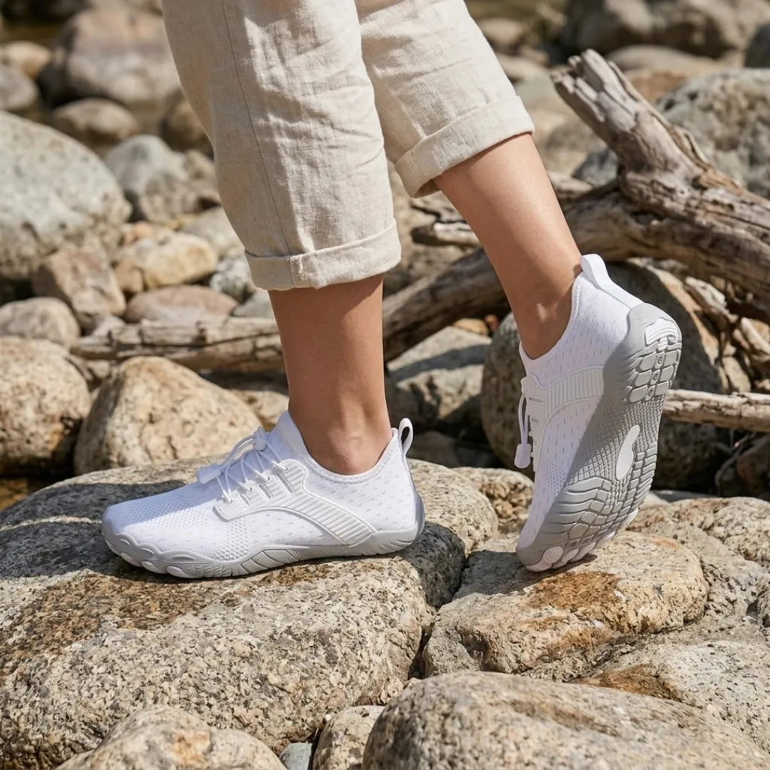 Model wearing white barefoot sneakers while stepping across sunlit rocks, one sole raised to show the gray tread.