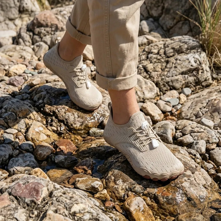 Model in rolled khaki pants wearing beige barefoot sneakers while walking over wet, rocky ground.