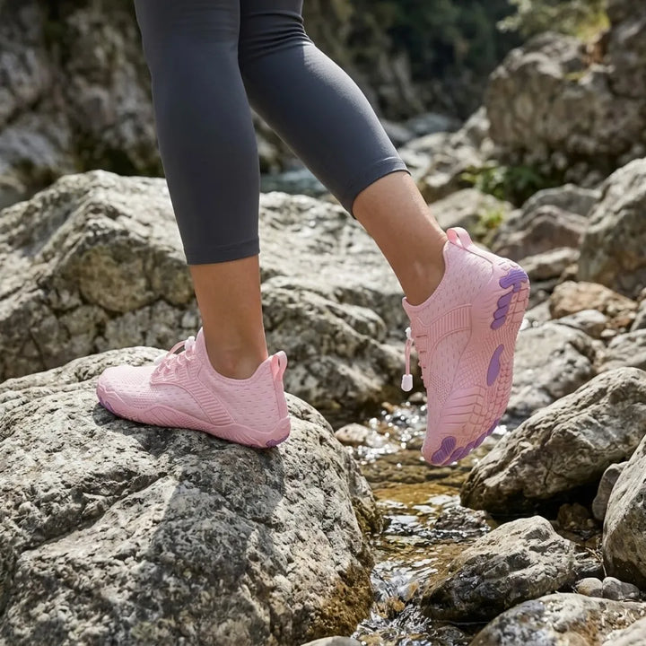 Woman in gray leggings stepping across creek rocks in pink sneakers, the purple-padded sole visible on the raised foot. 26