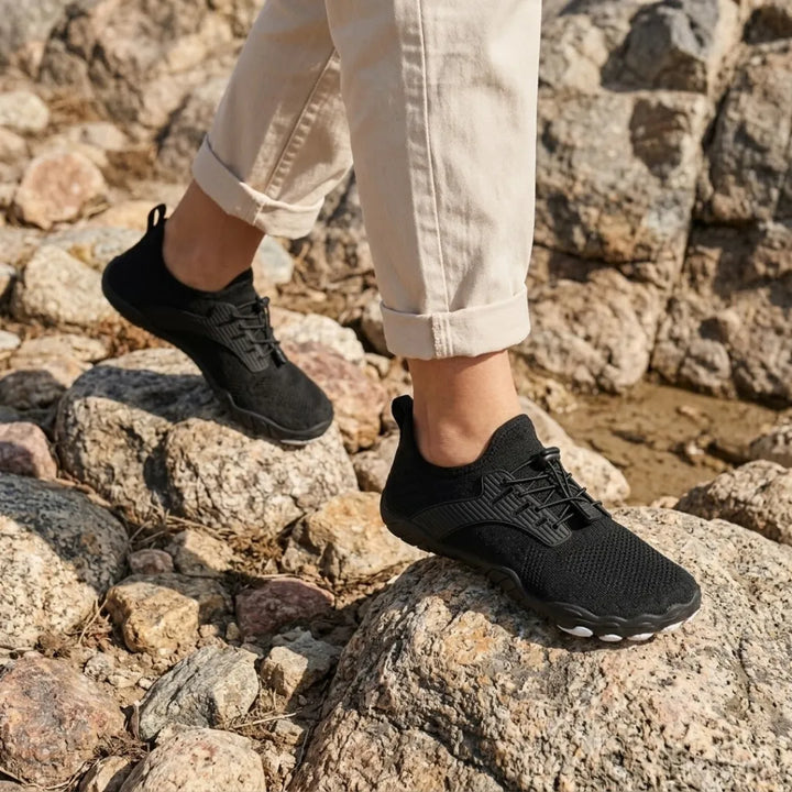 Person in rolled beige pants wearing black knit sneakers while walking over sunlit rocks. 33