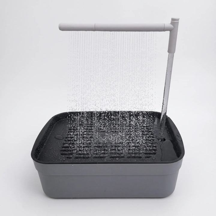 Gray bird bath with a dark slotted grate, a rain-style spray bar showering water down, and a vertical tube feeding it, on a white background. 1