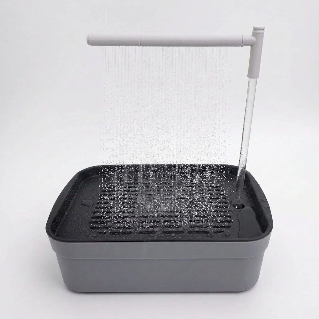 Gray rectangular bird bath on a plain white background with a slatted black tray and an overhead arm spraying water in thin streams. 1