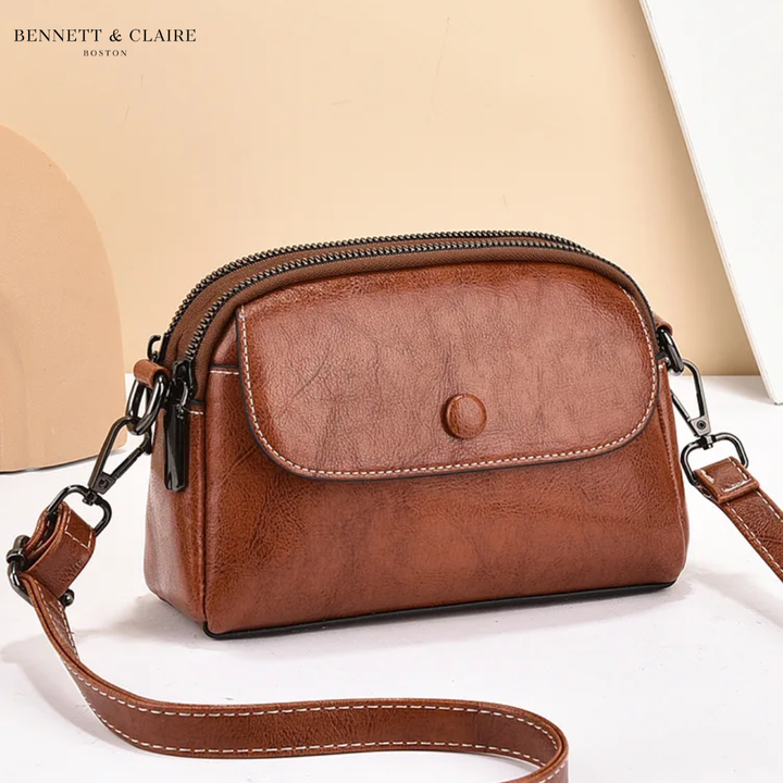 Tan-brown leather crossbody bag with front flap, snap button, contrast stitching and dark metal clips, shown angled against a peach backdrop. 1