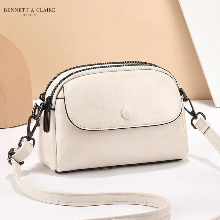 Cream white leather crossbody bag with front flap, snap button, double zipper and black hardware, angled against a peach backdrop. 4