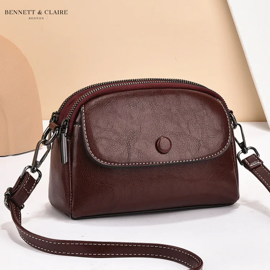Deep burgundy-brown leather crossbody bag with front flap, snap button and double zipper, strap curving across a white surface. 3