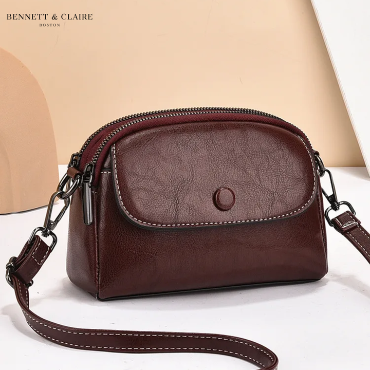 Deep burgundy-brown leather crossbody bag with front flap, snap button and double zipper, strap curving across a white surface. 3