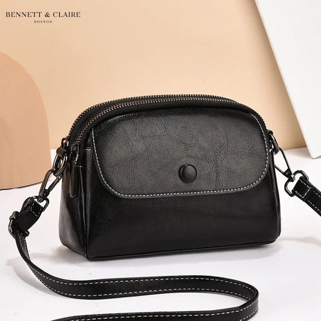 Black leather crossbody bag with front flap, snap button, white contrast stitching and double zipper top, angled against a peach backdrop. 2