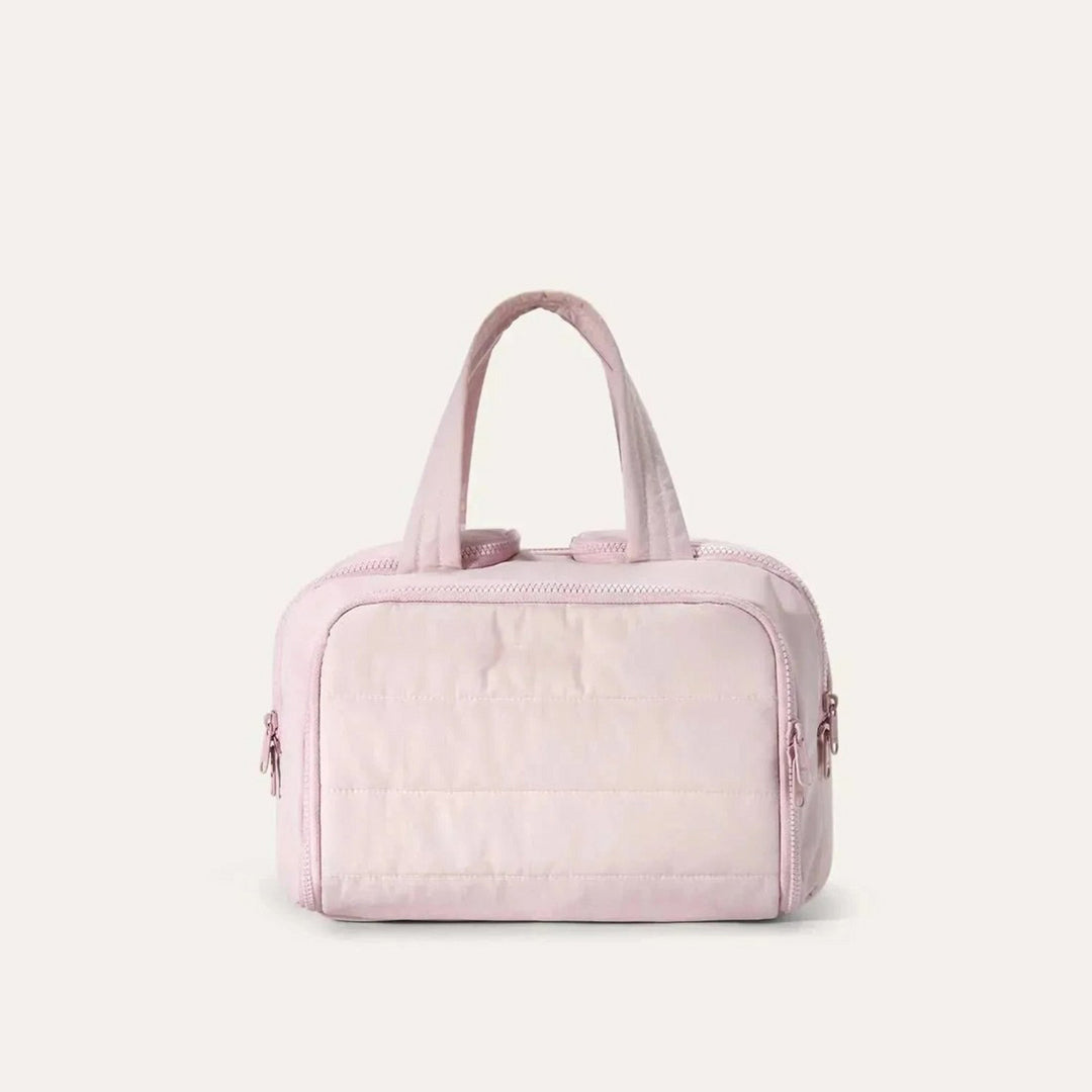 Pink quilted toiletry bag seen from the front, with two top handles and zippered pockets on the sides, on a plain light background. 1