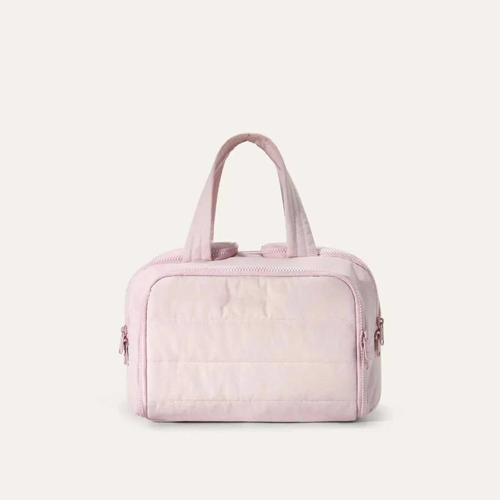 Pink quilted toiletry bag seen from the front, with two top handles and zippered pockets on the sides, on a plain light background. 1