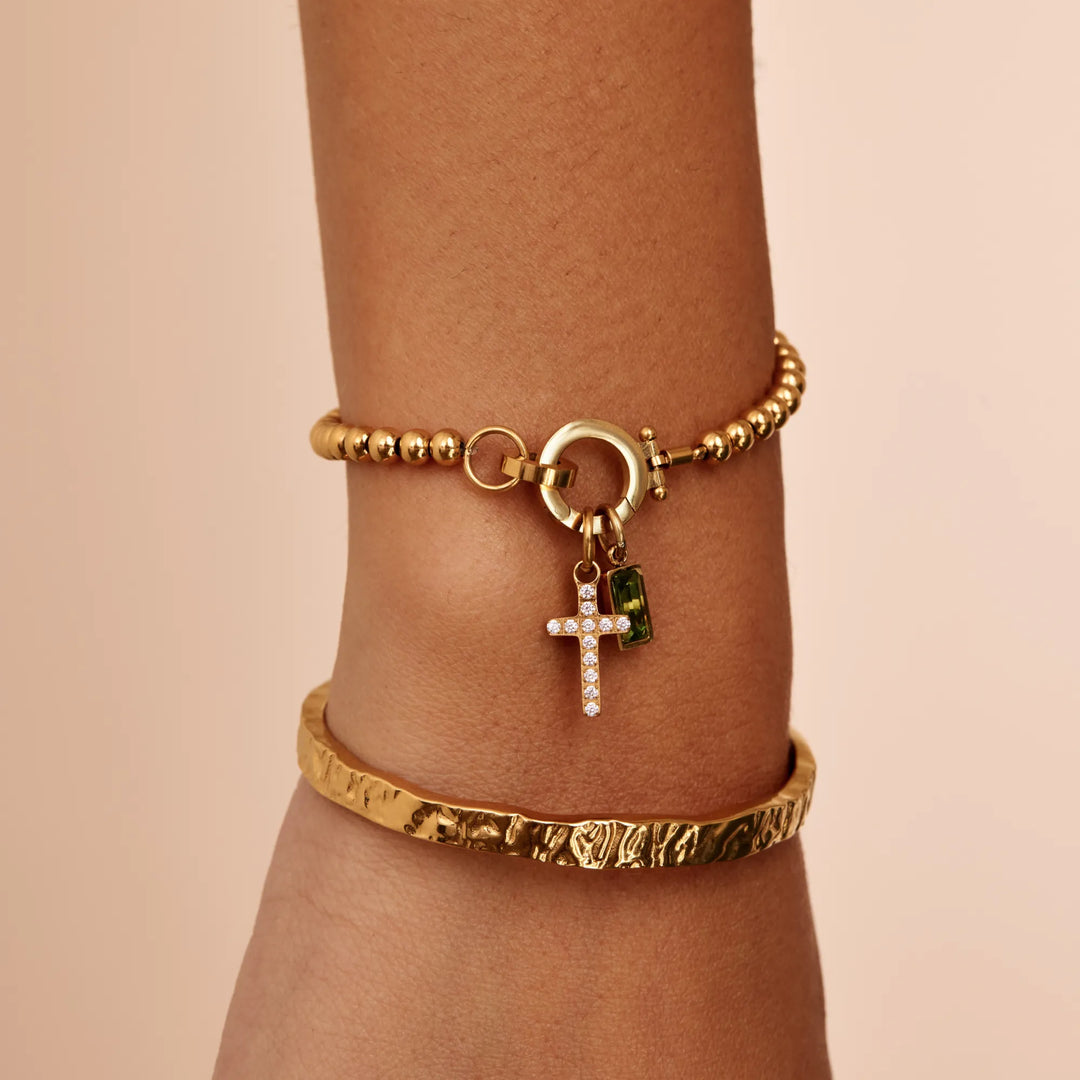 Gold-tone bead bracelet worn on a wrist with cross and green stone charms, stacked with a hammered gold cuff bangle.