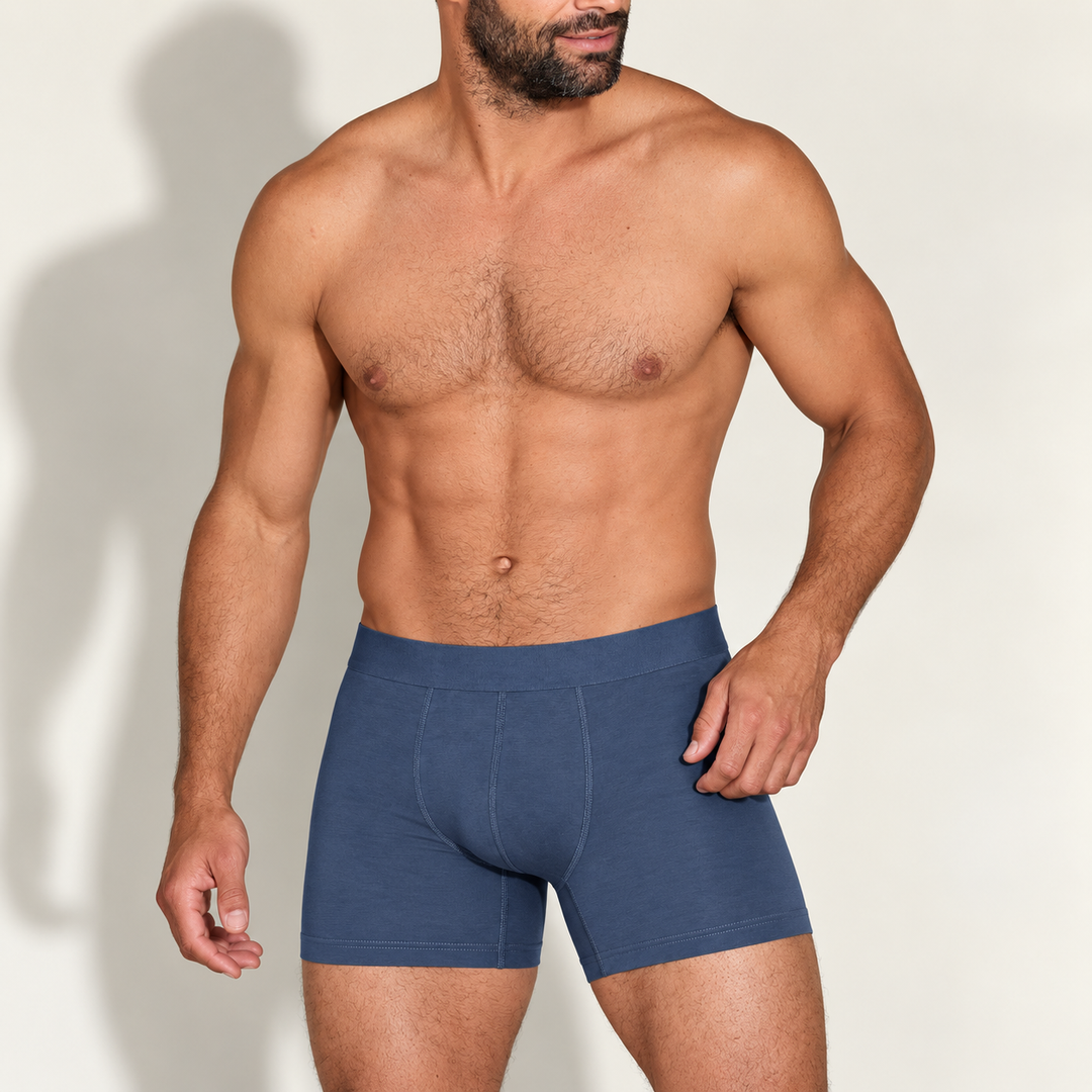 Bearded shirtless man wearing steel-blue modal boxer briefs with a wide waistband and front stitched seams, standing against a light wall. 1