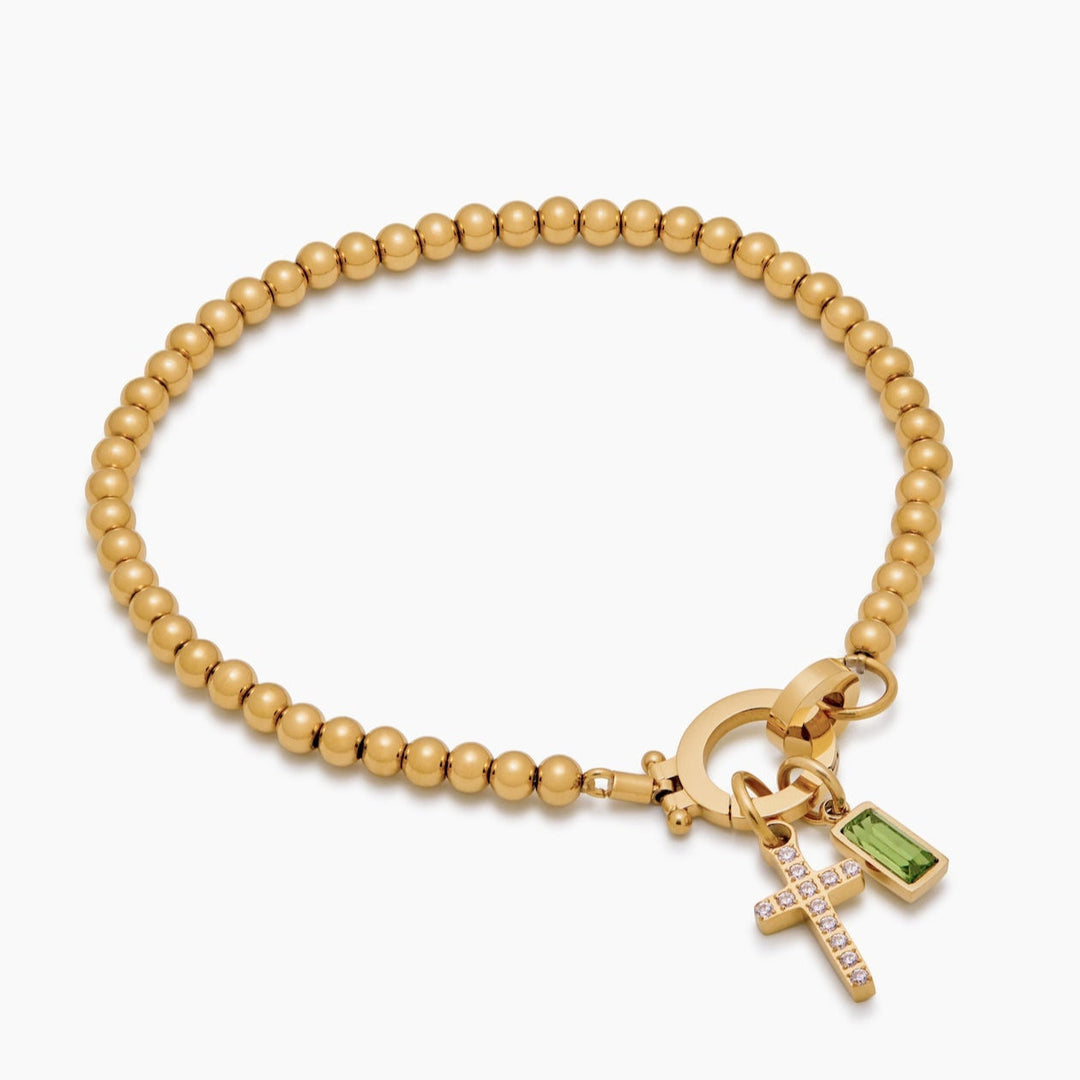 Gold-tone bead bracelet detail at an angle on white, with the cross charm and green baguette stone charm hanging from the round clasp.