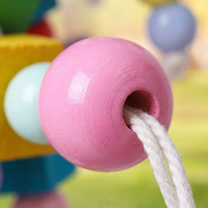 Close-up of a glossy pink wooden bead with white cotton rope threaded through it, with other colorful pieces blurred behind. 3