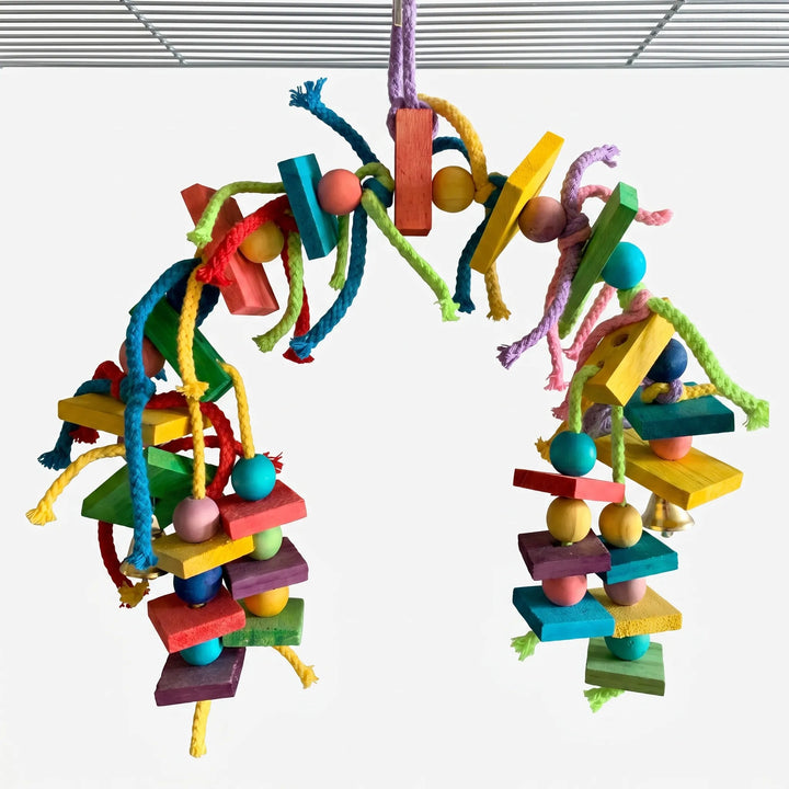 Arched bird toy of colored wooden blocks, beads, bells and knotted cotton ropes hanging from a cage roof against a white background. 1
