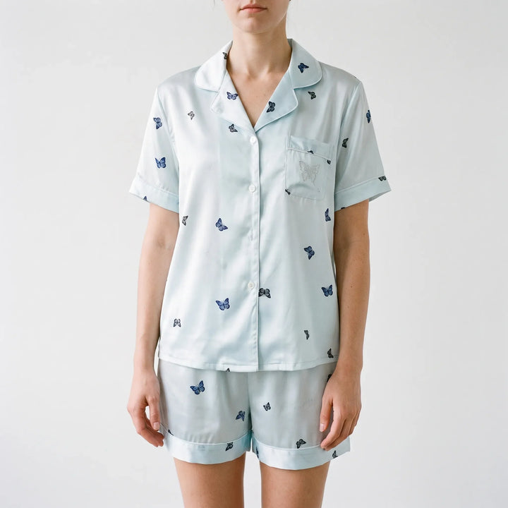 Front view on a model against a plain wall: short-sleeved button-up pajama top with butterfly print and a rhinestone butterfly on the chest pocket. 2