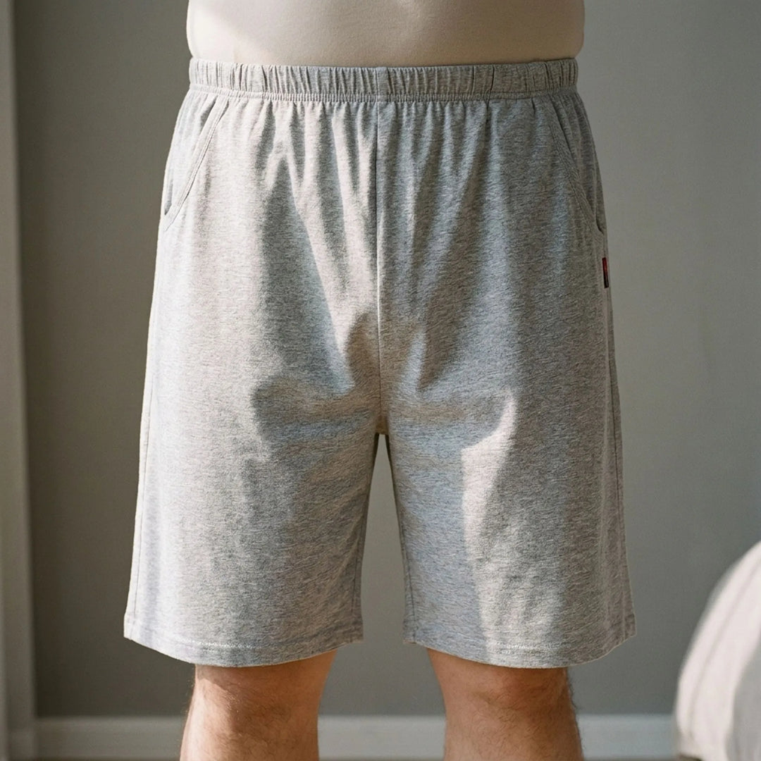 Man standing in a softly lit room wearing light heather gray jersey shorts with an elastic waistband and side pockets, front view. 1