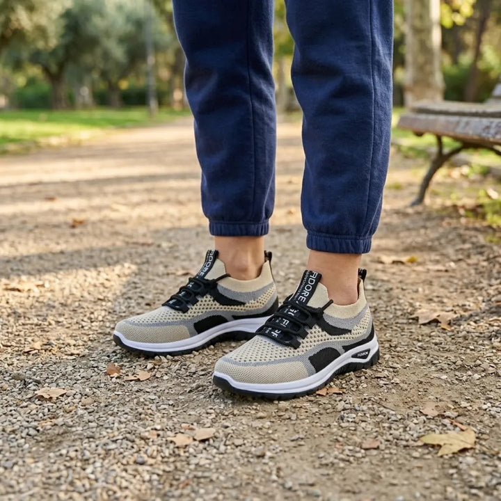 Person wearing beige knit mesh sneakers with black laces and white soles, paired with navy joggers, standing on a gravel park path. 38