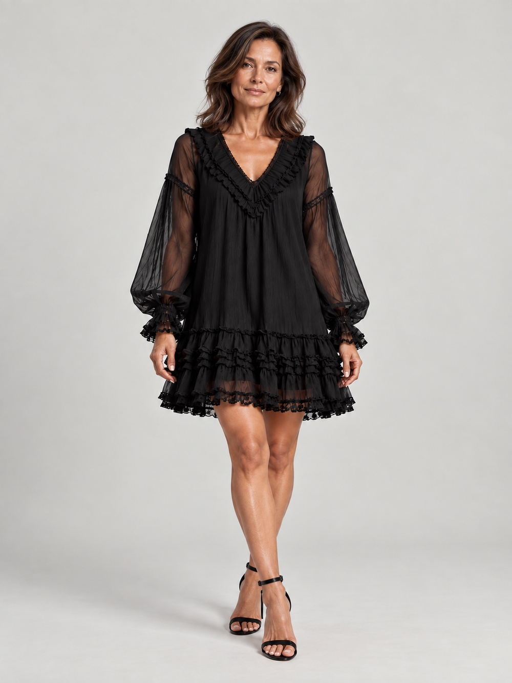 Full-length front view of a woman in the black ruffled mini dress with sheer sleeves, styled with black ankle-strap heels. 2