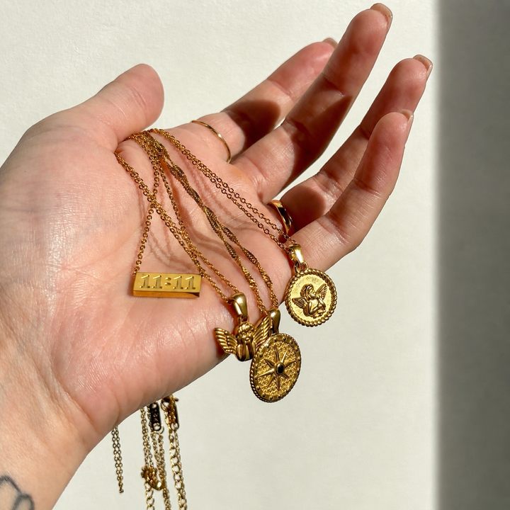Open palm holding several gold pendant necklaces, including the round cherub medallion, a star disc, a small winged figure and a bar. 3