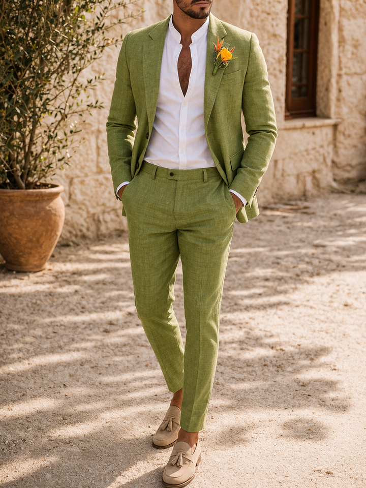Model in the olive green suit with an open-collar white shirt, hands in pockets, outdoors by a stone wall.