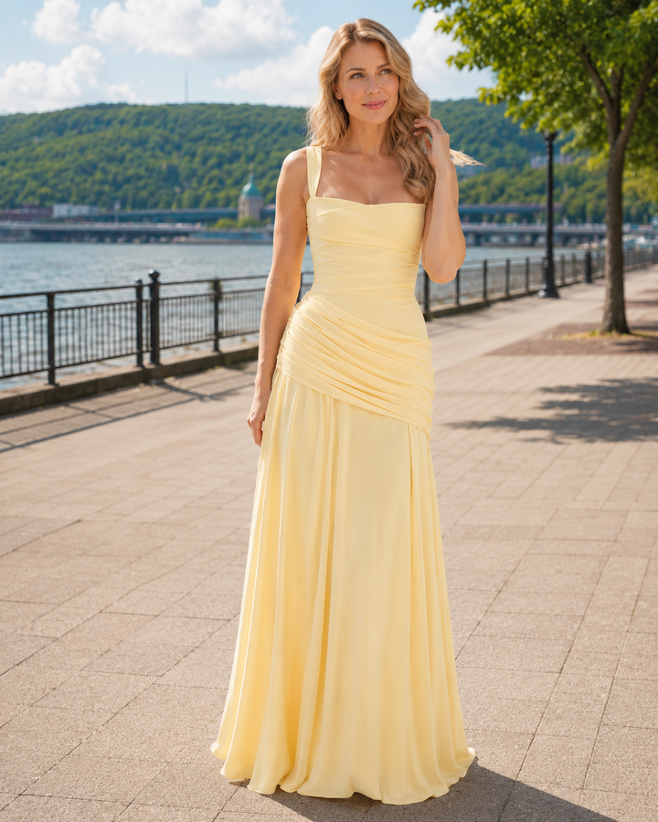 Woman in the pale yellow maxi dress standing on a sunny waterfront promenade, one hand in her wavy hair. 1