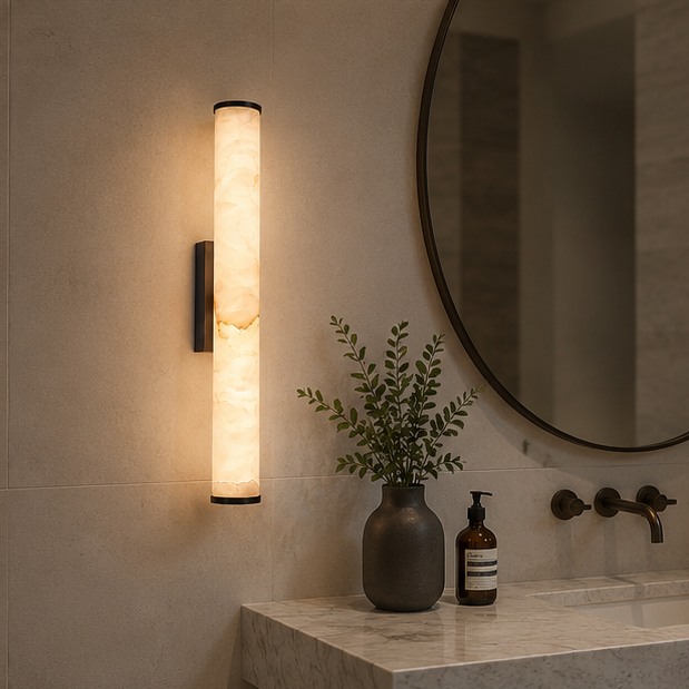 Black-finish marble sconce mounted on a tiled bathroom wall near a round mirror, a greenery vase, a soap bottle and a marble sink.